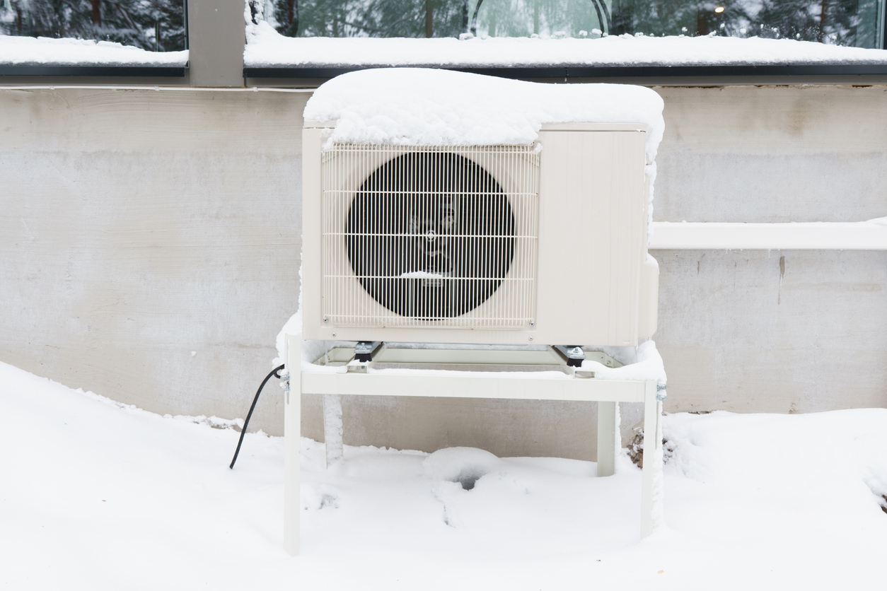 Outdoor heat pump on a metal stand beside a house, covered in fresh snow.